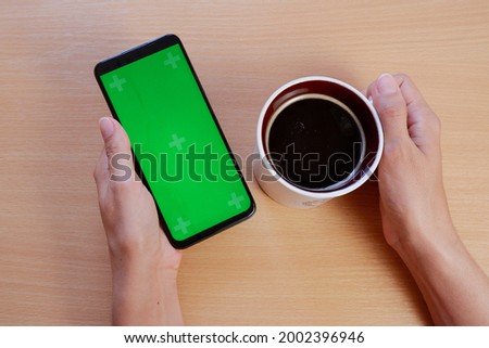 The hands of a young woman with a green-screen phone and a cup of coffee at the table. Chromakey for the layout.