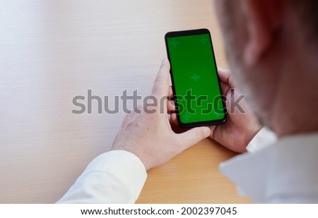A mock-up image of a hand man in a white shirt holding a black mobile phone with a blank green screen at a wooden table.