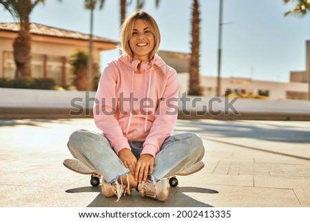 Young blonde skater girl smiling happy using headphones sitting on the skate