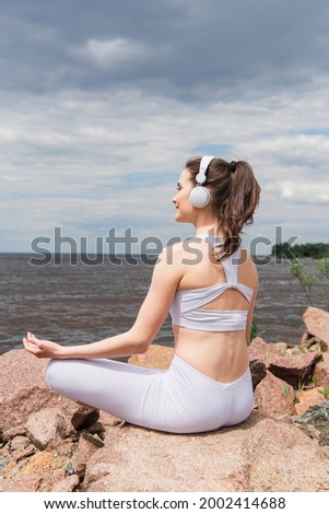 happy woman in headphones and sportswear sitting in lotus pose while meditating near sea