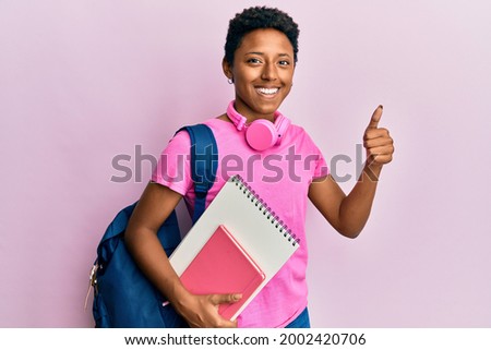 Young african american girl wearing school bag and holding books smiling happy and positive, thumb up doing excellent and approval sign 