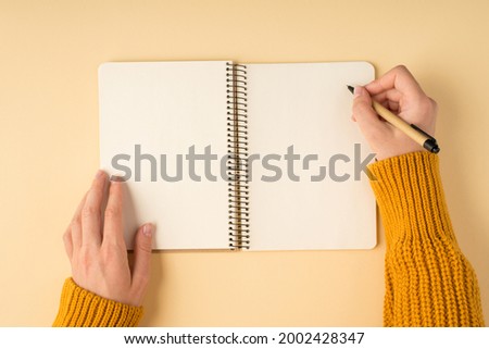 First person top view photo of female hands in yellow knitted sweater writing in spiral notebook on isolated pastel orange background with blank space