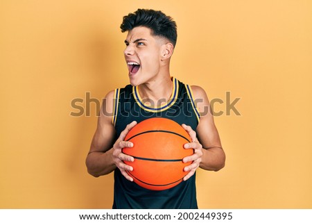 Young hispanic man holding basketball ball angry and mad screaming frustrated and furious, shouting with anger. rage and aggressive concept. 
