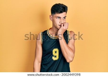 Young hispanic man wearing basketball uniform looking stressed and nervous with hands on mouth biting nails. anxiety problem. 