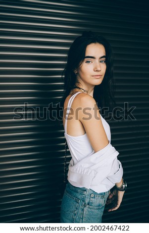 Portrait happy girl with long dark hair wearing white top and blue jeans standing over brown blinds background looking at camera.