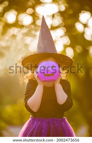Happy Halloween. Portrait of a smiling girl in a witch hat with a purple bucket of pumpkin sweets in the autumn forest. Cheerful children in carnival costumes in nature.