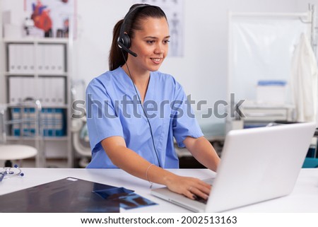 Medical receptionist wearing headset with microphone in private hospital typing on laptop Health care physician sitting at desk using computer in modern clinic looking at monitor. Royalty-Free Stock Photo #2002513163