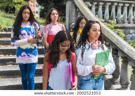 Close-up of two Latina teenage student friends back to school with backpacks, with three more girlfriends out of focus in the background