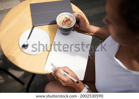 High angle view of a woman writing on a notebook sitting at a table with a coffee in her hand. Freelance, business and outdoor work concepts