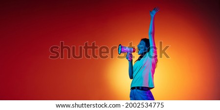 Shouting. Young stylish woman with loudspeaker isolated on orange studio background in neon light. Female model in casual style. Concept of human emotions, facial expression, youth. Copy space for ad.