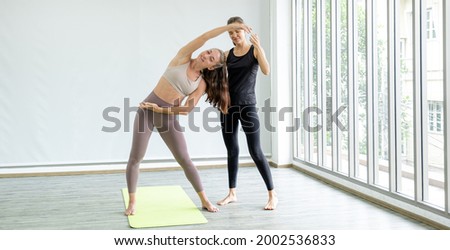 A young caucasian woman is teaching yoga stretching pose to her friend at home. Royalty-Free Stock Photo #2002536833
