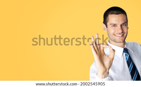 Portrait of smiling businessman in white shirt and tie, showing thumb up like hand sign gesture, isolated over orange yellow colour background. Happy confident man gesturing. Business success concept.