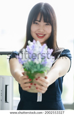 Vertical portrait shot of attractive smiling young Asian woman with long straight hairstyle in casual blue clothes holding the purple and white flowers while looking at camera in the studio