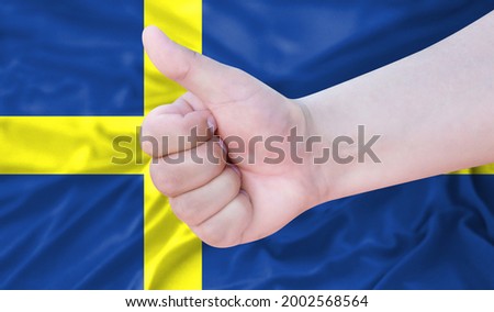 Hand makes a thumbs up sign on the background of the flag of Sweden. like, good, positive