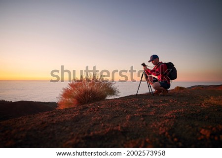 Young man during photographing landscape on top of hill above clouds. Hobby photographer waiting for beautiful sunset. Tenerife, Canary Islands, Spain.
