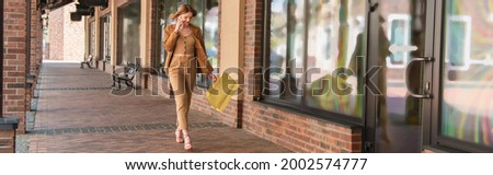 Cheerful customer with shopping bags talking on mobile phone while walking near building, banner