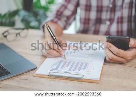 Close-up businessman holding a pen, smartphone and pointing at financial graph checking business report on wooden desk with computer laptop besided at home. Stock photo