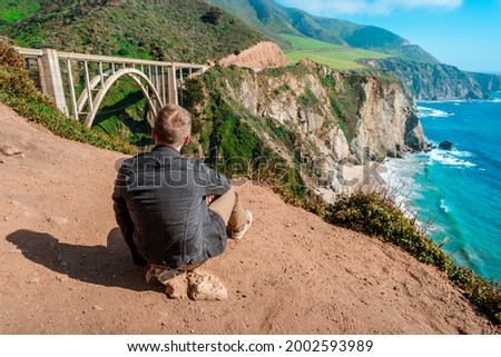 A young man in a denim jacket on a slope overlooking the Bixby Creek Bridge on the Big Sur coast in California