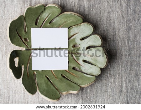 Summer stationery still life. Blank business card mockup on ceramic plate of monstera sheet. Gray linen tablecloth fabric background. Branding concept. Mediterranean design. Styled flat lay