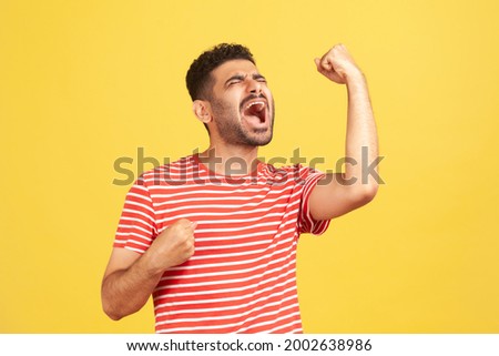 Extremely excited overjoyed man with beard in striped t-shirt shouting making yes gesture, amazed with his victory, triumph. Indoor studio shot isolated on yellow background Royalty-Free Stock Photo #2002638986
