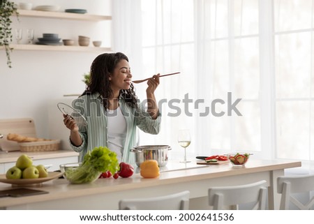 Diet, healthy lifestyle. Cook at home, household and nutrition. Glad young african american woman cooks soup and tastes food in modern kitchen interior with bright vegetables, on window background Royalty-Free Stock Photo #2002641536