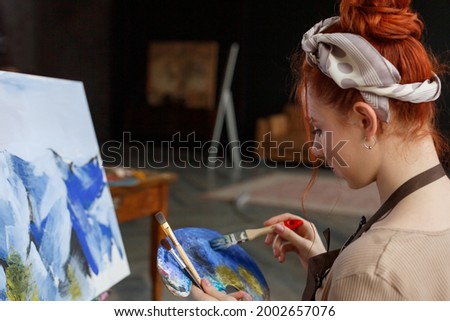 Talented female artist working on a painting in a dark creative studio. Beautiful creative girl sits in a workplace near a painted picture snowy mountains