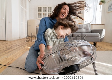 Playful single mother bonding with son excited playing together at home with big fan blowing cool wind in living room. Mum and child enjoy time together at conditioning ventilator during summer heat Royalty-Free Stock Photo #2002692806
