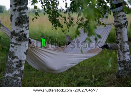 A man who rests or works remotely in the countryside or in the garden, lies in a hammock under the birches and uses or looks at a smartphone with a green screen layout.