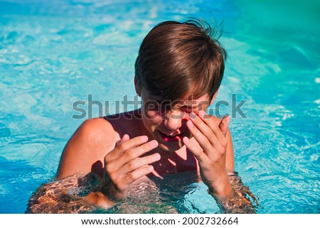 The teenager hides his face with his hands in the pool, wiping water from his eyes while swimming in the pool. Eye irritation from chlorinated water Royalty-Free Stock Photo #2002732664