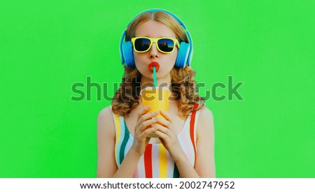 Summer portrait of stylish young woman listening to music in headphones and drink fresh juice on green background