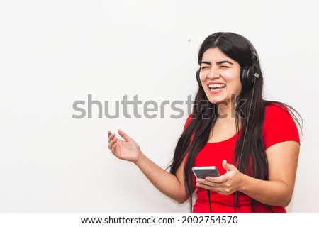 pretty girl, hispanic latina, white skin, black hair, wearing a red flannel, is smiling happily with her phone in her hand and is wearing large black headphones and is on a white background.