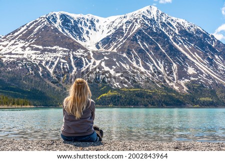 Blonde female woman sitting beside lake in northern Canada with huge snow capped mountains in background with scenic, amazing view for tourism, tourist, road trip. 