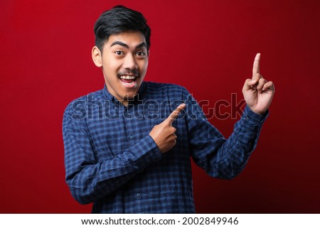 Young handsome asian man wearing casual shirt over red background with a big smile on face, pointing with hand finger to the side looking at the camera.