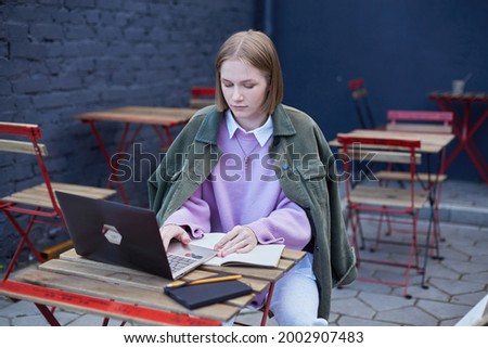 Young woman freelancer in glasses sits on cafe terrace at table with black laptop and types under bright sunlight. Female student distant learning foreign languages and working distantly. Royalty-Free Stock Photo #2002907483