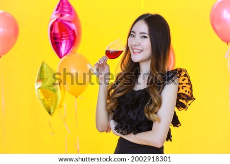 Portrait of beautiful smiling young asian woman with  glass of red wine with yellow background and party balloon.