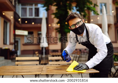 Disinfecting to prevent COVID-19. Waiter in protective face mask and gloves cleaning table with disinfectant spray and Microfiber cloth in cafe  in quarantine city.