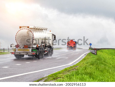 Two tanker trucks for transporting petroleum products drive on the highway with their headlights on in cloudy weather and rain in summer. Gasoline transportation concept, dangerous cargo Royalty-Free Stock Photo #2002956113