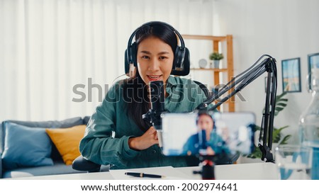 Teenage Asia girl influencer use microphone wear headphone record content with smart phone for online audience listen at house. Student female podcaster make audio podcast from her home studio. Royalty-Free Stock Photo #2002974641