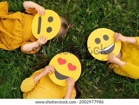 Three children lying on the grass are holding cardboard emoticons with different emotions in their hands: a sad, smiling happy smile, a loving smile with hearts instead of eyes.  World Emoji Day Royalty-Free Stock Photo #2002977986