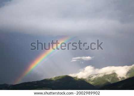 Rainbow on a dark grey sky background with some rays of sun shining through the clouds. On the bottom of the picture are some hills behind white clouds.