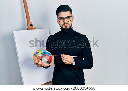 Handsome man with beard holding palette and brush close to easel stand puffing cheeks with funny face. mouth inflated with air, catching air. 