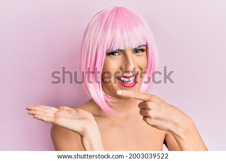 Young man wearing woman make up wearing pink wig amazed and smiling to the camera while presenting with hand and pointing with finger. 