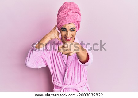 Young man wearing woman make up wearing shower towel on head and bathrobe smiling doing talking on the telephone gesture and pointing to you. call me. 