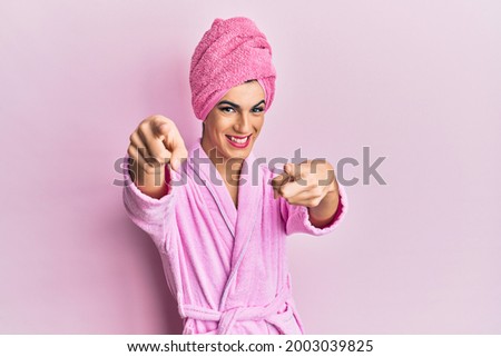 Young man wearing woman make up wearing shower towel on head and bathrobe pointing to you and the camera with fingers, smiling positive and cheerful 