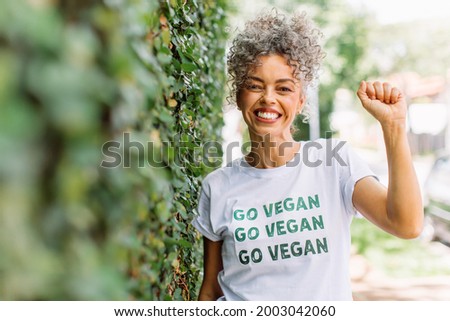 Vegan activist smiling cheerfully while standing alone outdoors. Happy mature woman advocating for veganism while wearing a shirt with the words "GO VEGAN" written on it. Royalty-Free Stock Photo #2003042060