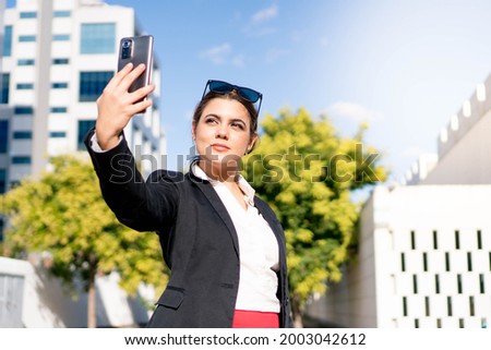 Business woman dressed in a suit, red skirt and sunglasses, taking a photo outdoors in her workspace. Successful young worker making a video call with buildings in the background in a modern space.