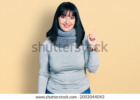 Young hispanic plus size woman wearing winter scarf with a big smile on face, pointing with hand finger to the side looking at the camera. 