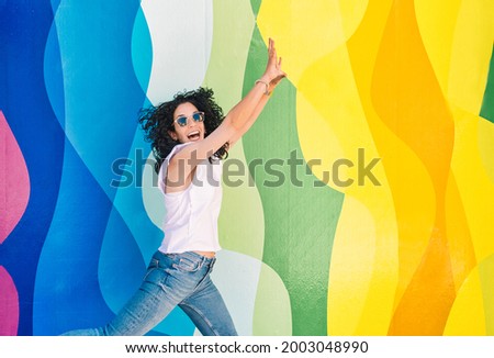 Young brunette woman with curly hair, jeans and sunglasses has fun while jumping on a colorful wall. leisure and free time concept