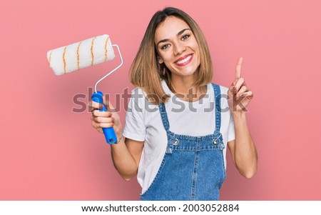 Young blonde girl holding roller painter surprised with an idea or question pointing finger with happy face, number one 