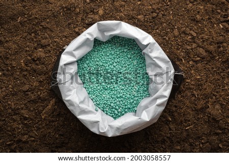 Opened plastic bag with green complex fertiliser granules on dark soil background. Closeup. Product for root feeding of vegetables, flowers and plants.  Royalty-Free Stock Photo #2003058557